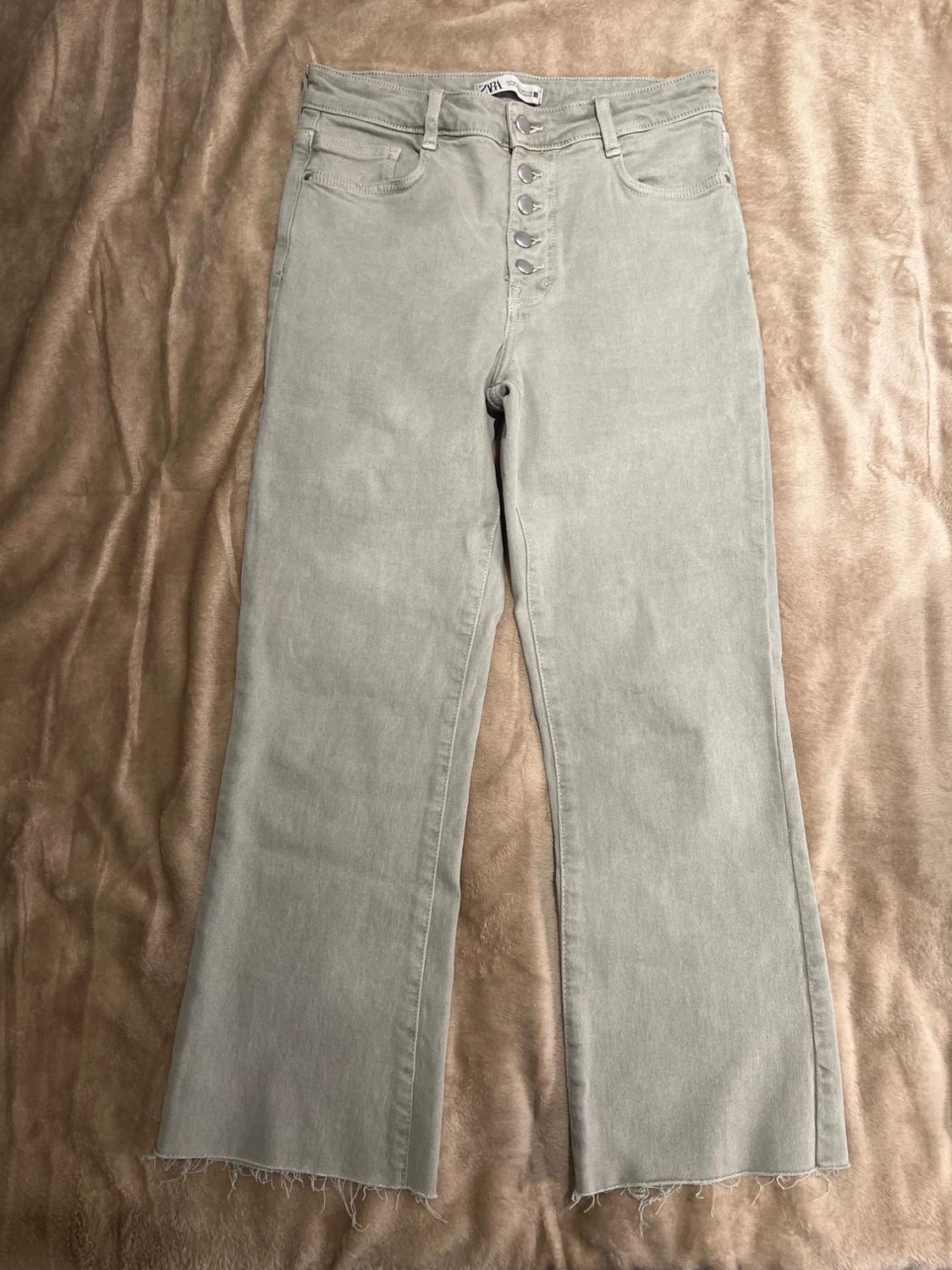 Grey denim trousers with visible buttons, frayed hem, on a beige fabric surface.