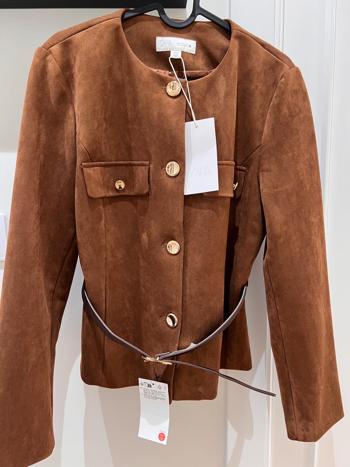 Brown suede jacket with gold buttons and a thin belt, hanging on a hanger. Visible label with text "ZARA".