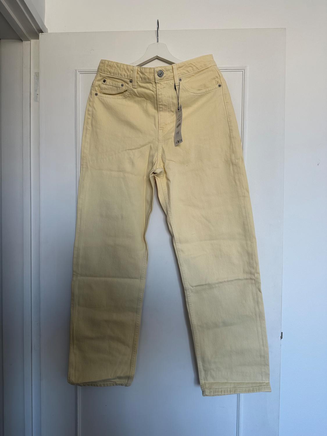 Yellow trousers hanging on a white hanger over a white door. Label visible on the front pocket.