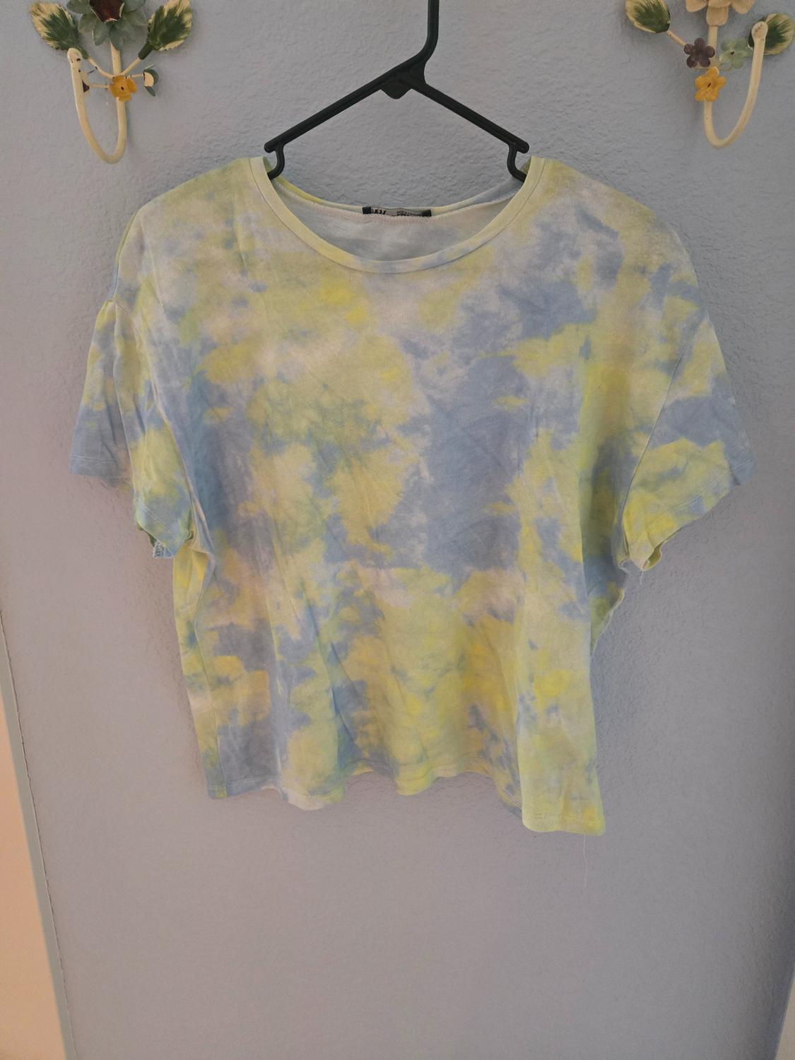 Short-sleeved T-shirt with a blue and yellow tie-dye design, hanging on a black hanger against a grey wall.