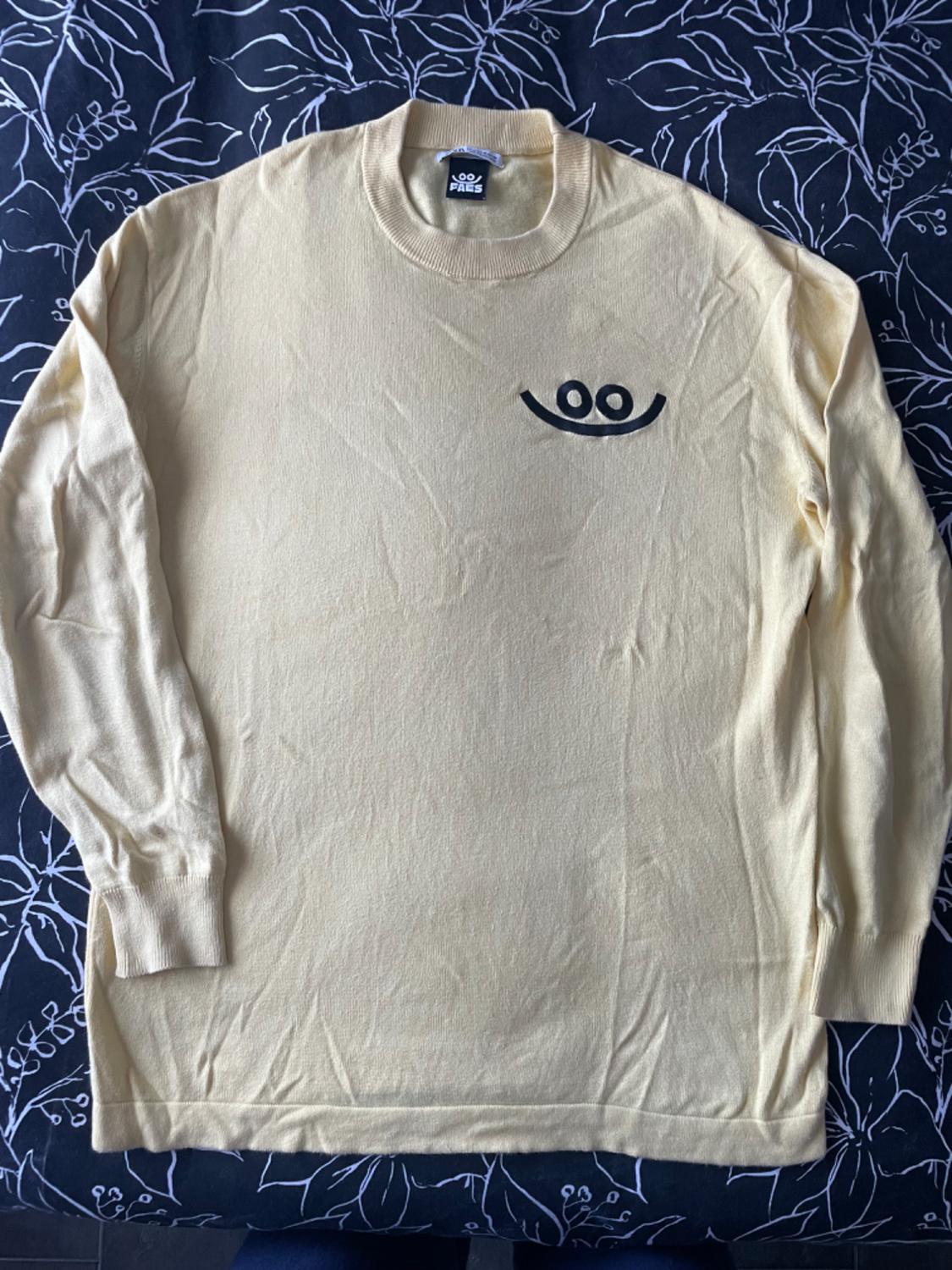 Beige long-sleeved jumper with eye and smile design on the chest. Black background with white leaf pattern. "PAES" label.