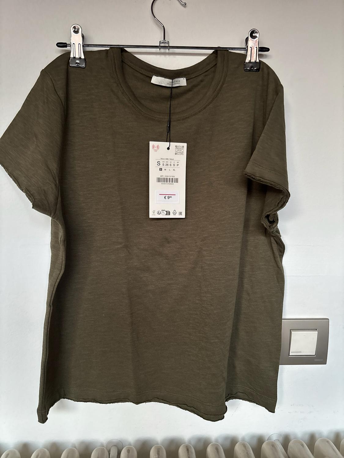 Olive-green short-sleeved T-shirt, frayed edges, hanging on a hanger against a white wall. Label with text: S 26 S S P, S M L XL, € 9.95.