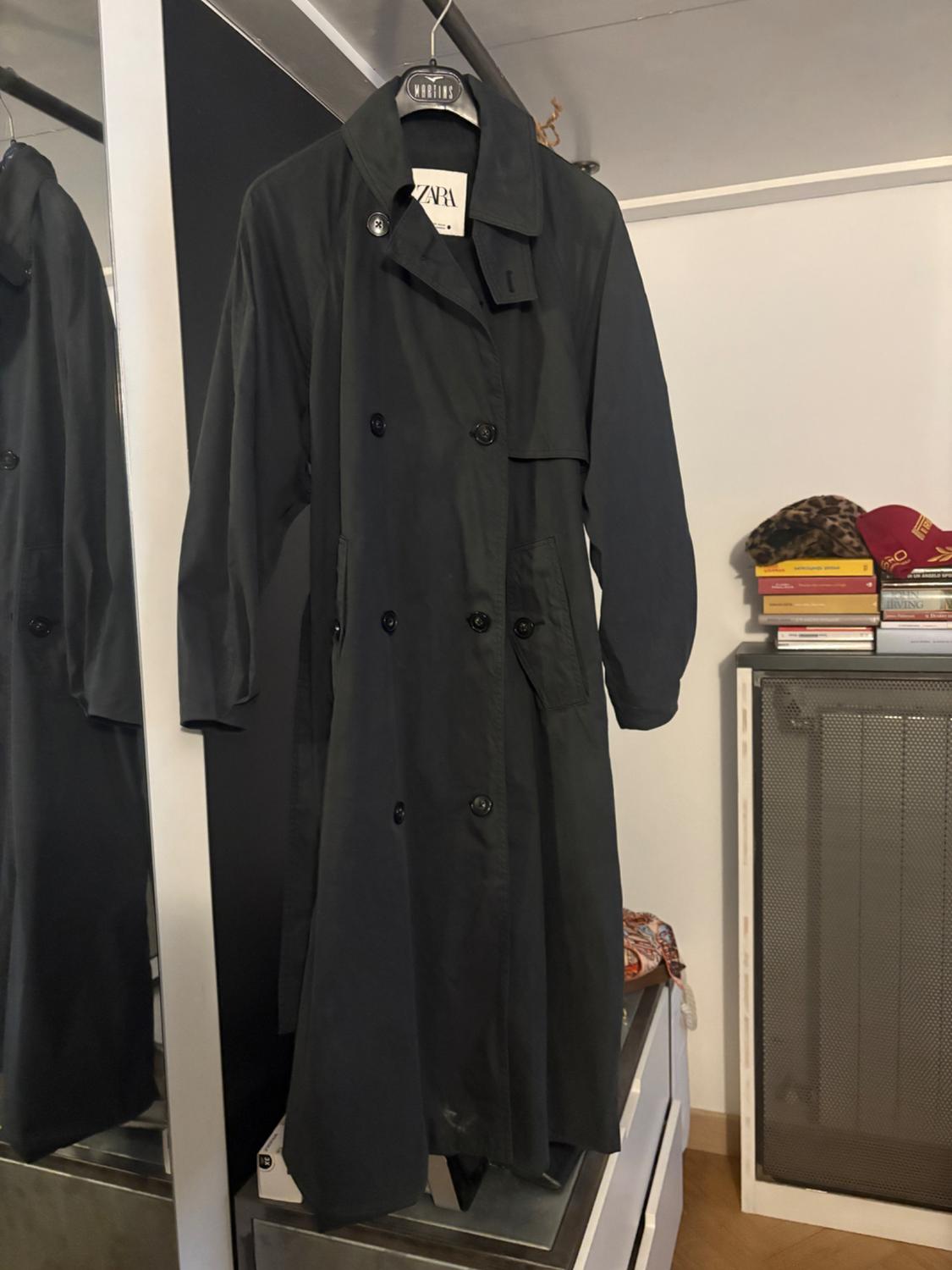 Black coat hanging on a hanger with a visible tag. Reflection in a mirror, background with books and a cap on a piece of furniture.