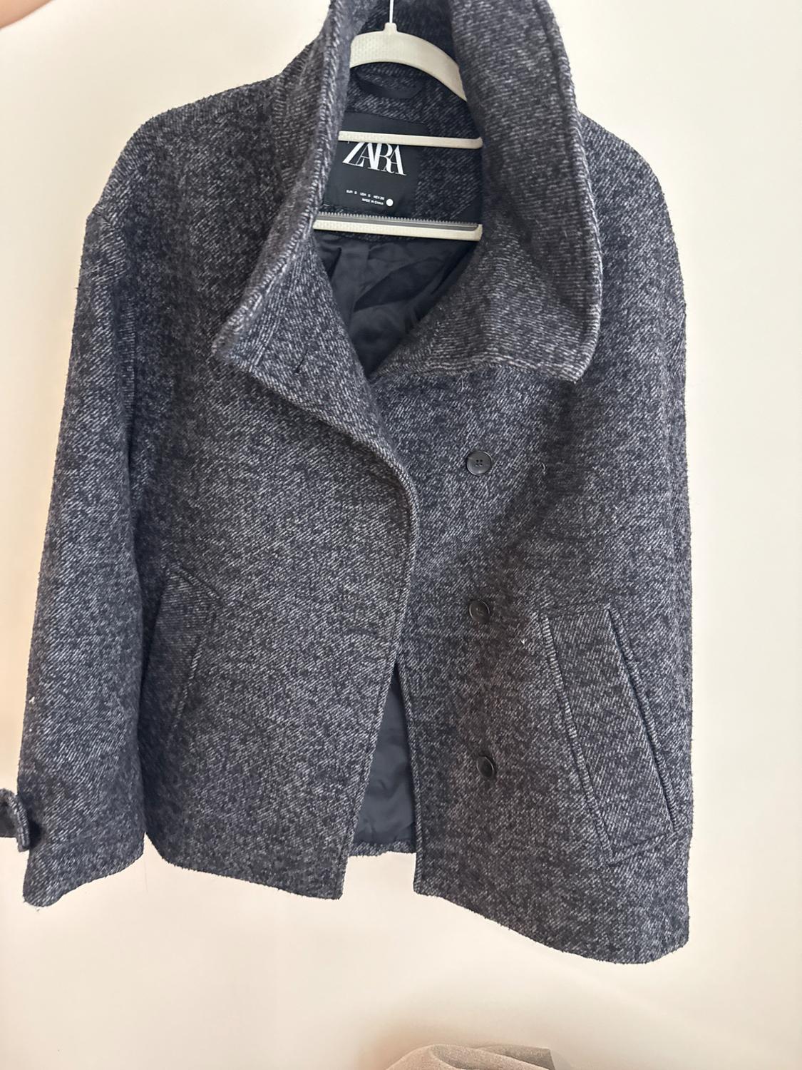Short marled dark gray coat with a wide collar and buttons, hanging on a white hanger against a light background. ZARA text on the label.