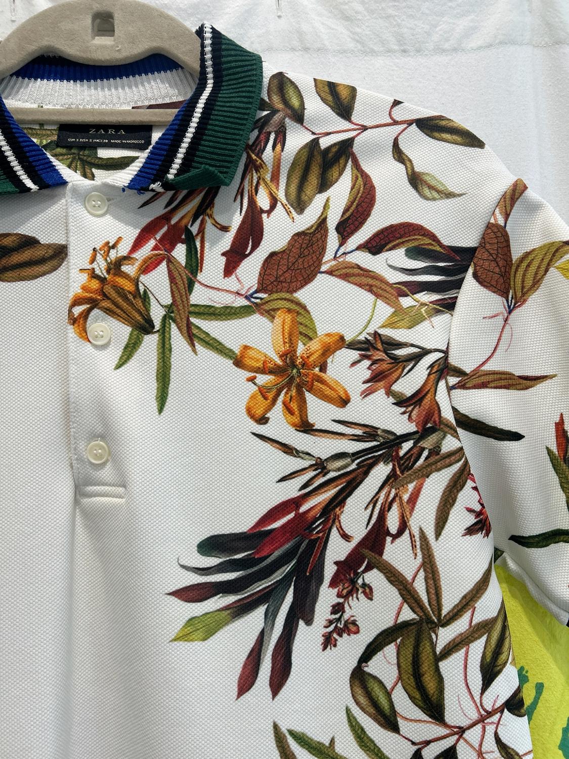 White polo shirt with floral print and a green, blue, and white striped collar. Inner label with ZARA text. Hanging on a hanger.