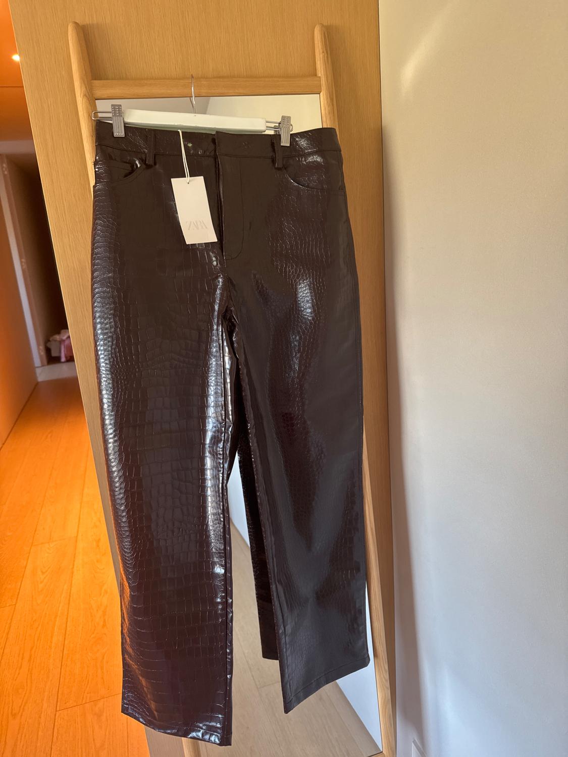 Shiny textured brown trousers hung on a wooden hanger. Visible tag with the text "ZARA". Light wood background.