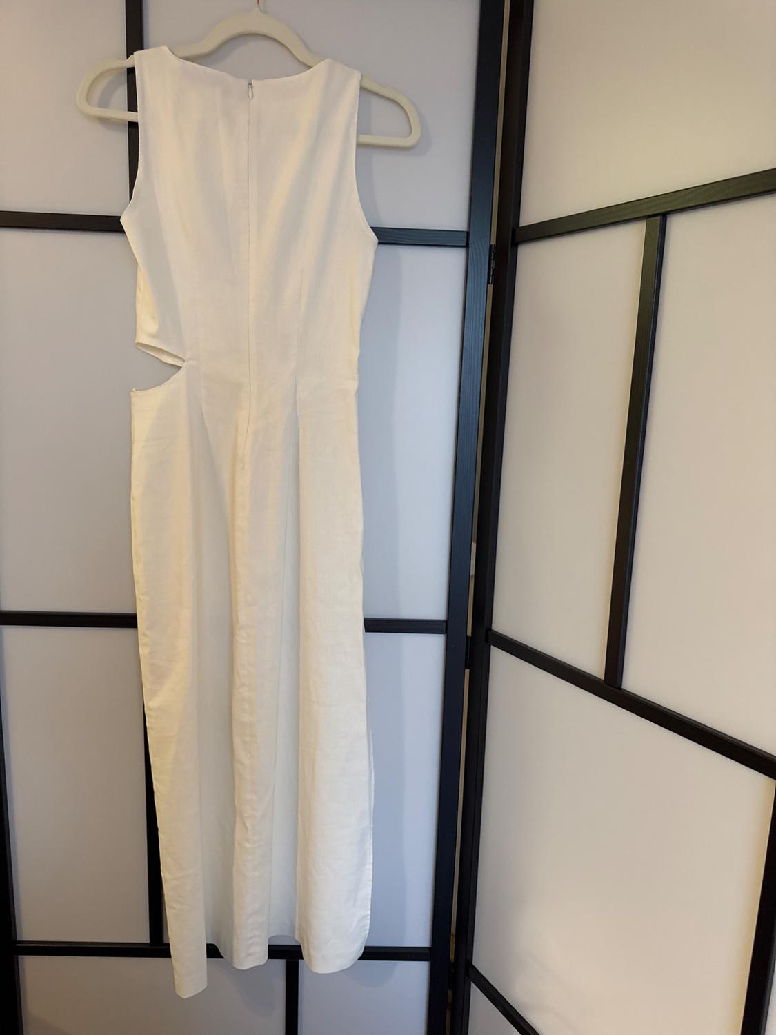 Sleeveless white dress hanging on a hanger, with a side-cut design, against a wooden panel and glass background.