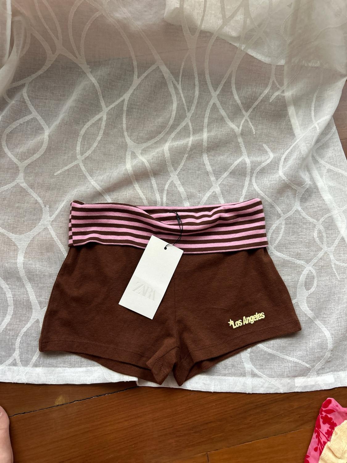 Brown shorts with a pink striped waistband and ‘Los Angeles’ text. ZARA label. Image on white fabric and a wooden floor.