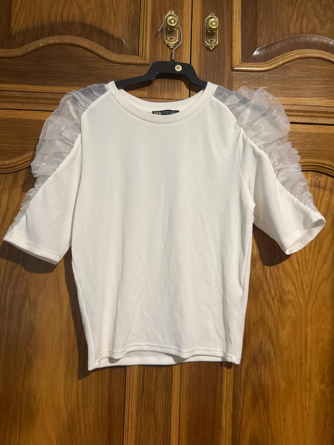 White short-sleeved blouse with sheer ruffles on the shoulders, hanging on a black hanger over a wooden door.