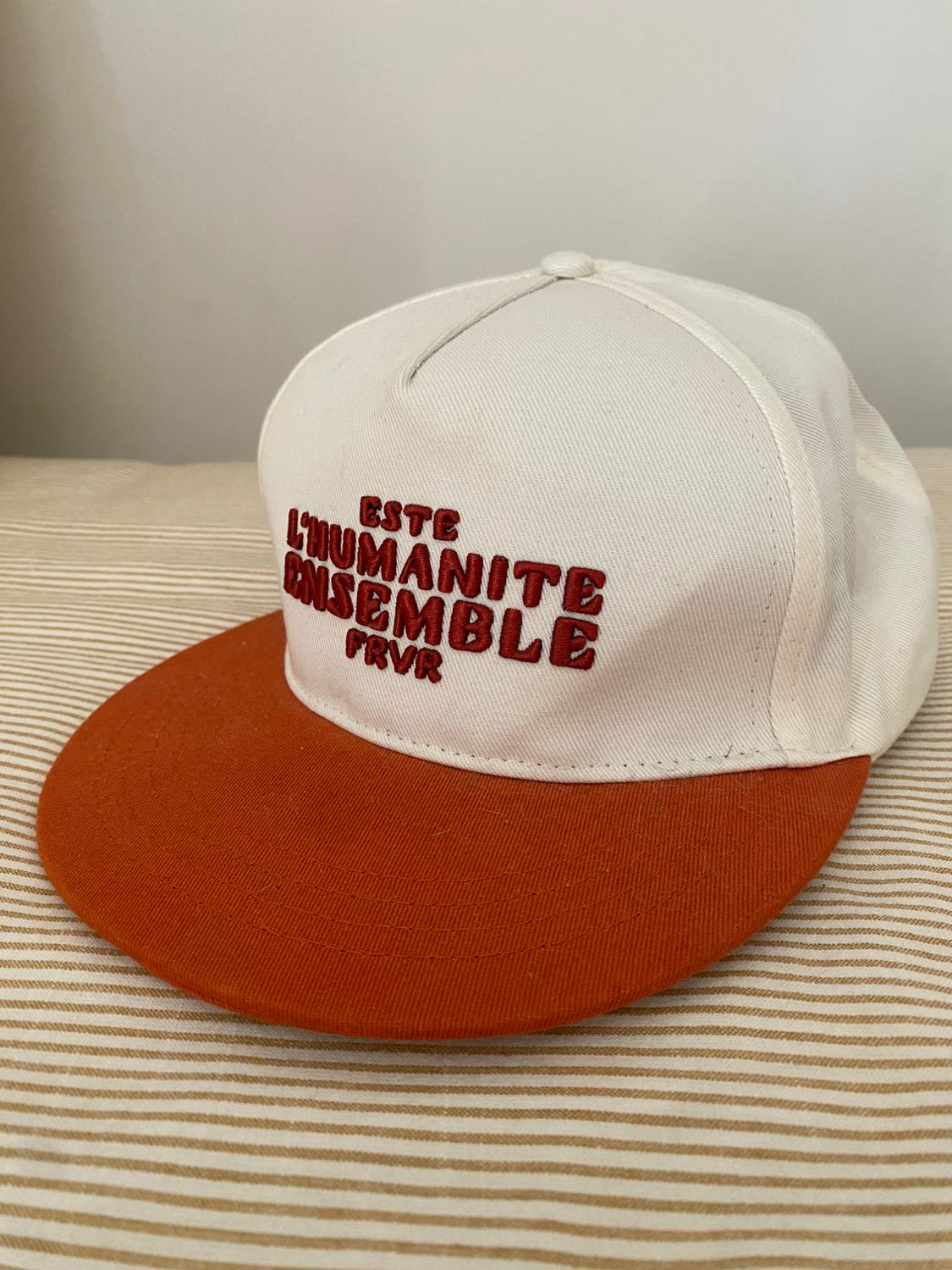 White cap with orange peak on striped fabric. Text on cap: "ESTE L'HUMANITE ENSEMBLE FRVR" in red letters.