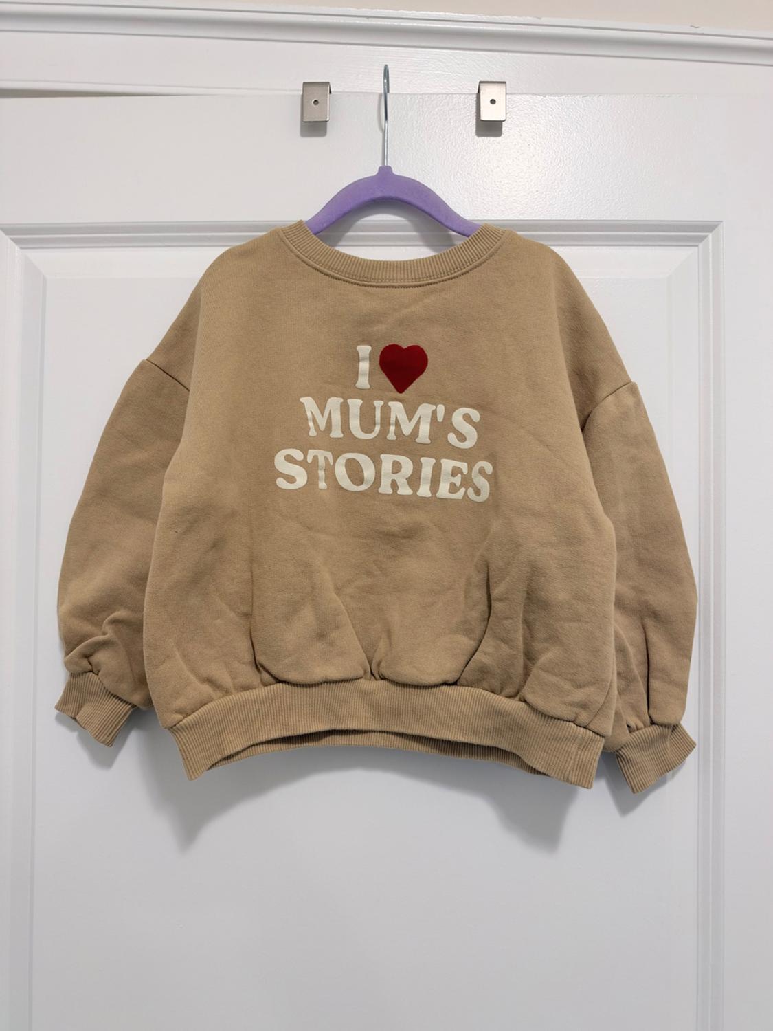 Beige sweatshirt hanging on a purple hanger on a white door. It features a red heart and the white text "I MUM'S STORIES".