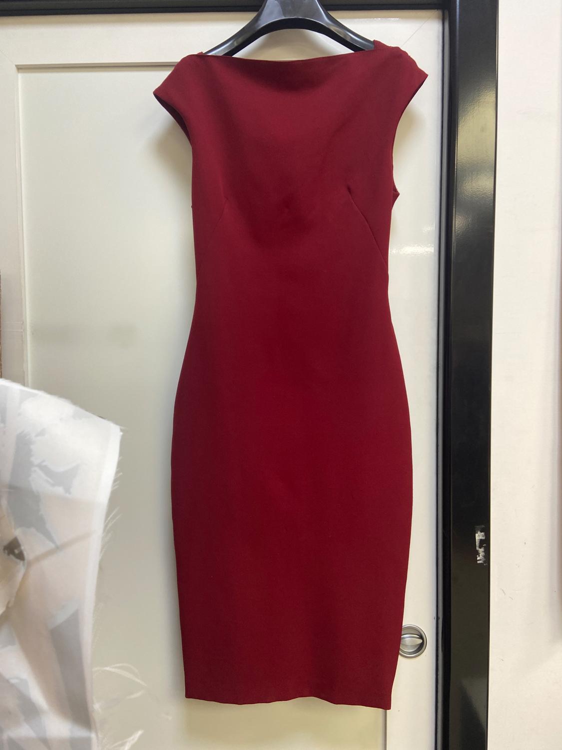 Dark red fitted dress, sleeveless and with a boat neckline, hanging on a black hanger in front of a white door.