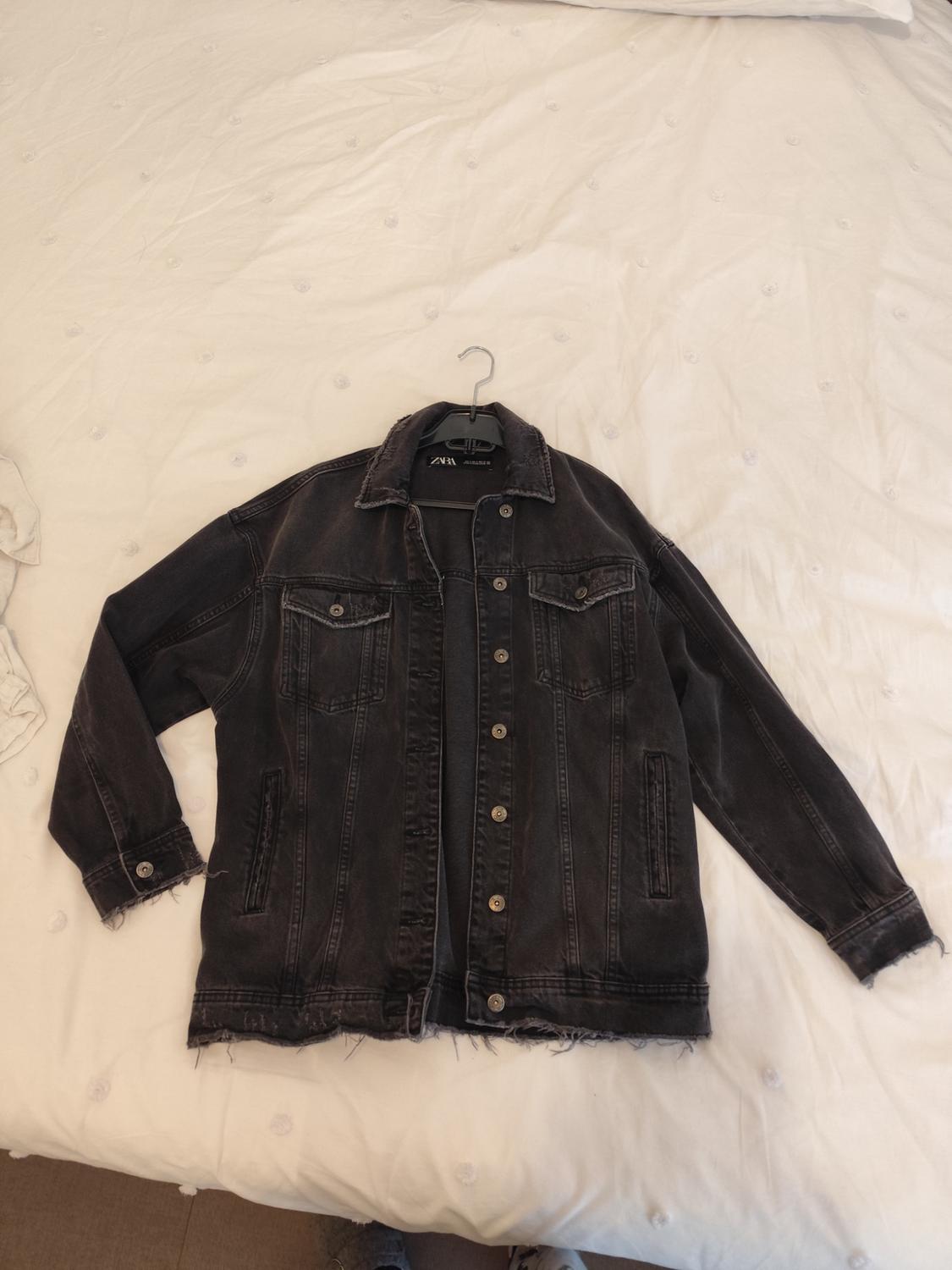 Worn black denim jacket on a white bed. The jacket is on a hanger with the label visible.
