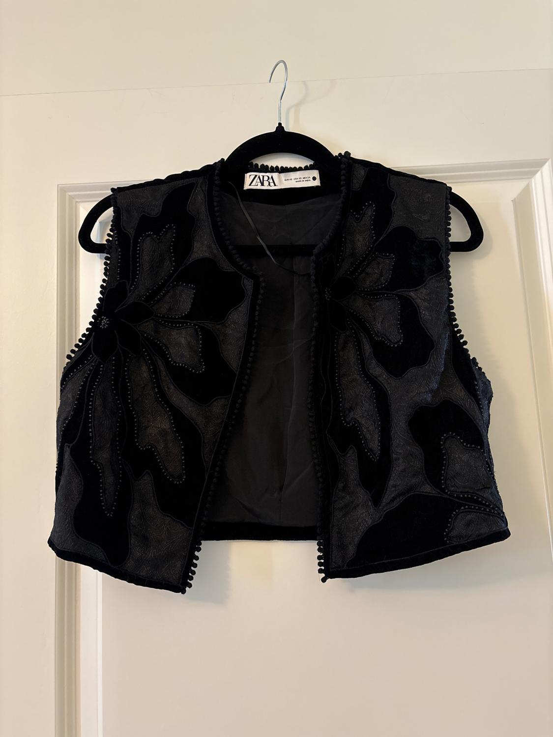 Black waistcoat with a raised floral design, hanging on a black hanger. Label visible with the text "ZARA". White door background.