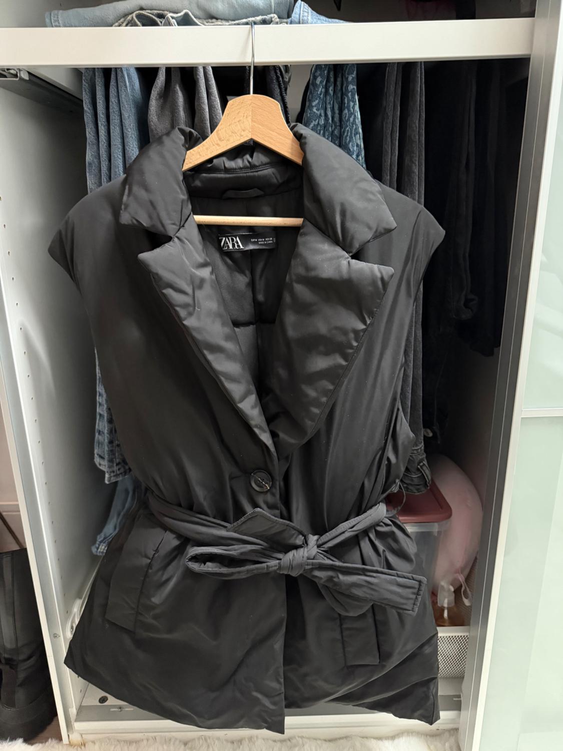 Black quilted gilet with a belt hanging in a wardrobe, surrounded by clothes in dark and light shades. Visible tag: Zara.