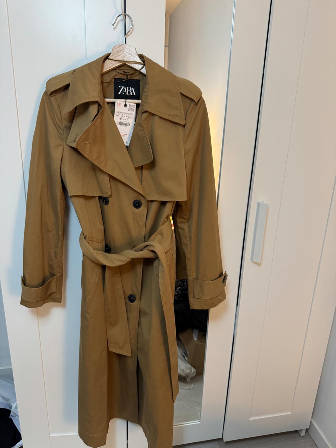 Brown trench coat hanging on a hanger in front of a white wardrobe. Visible label with Zara logo and XS size details.