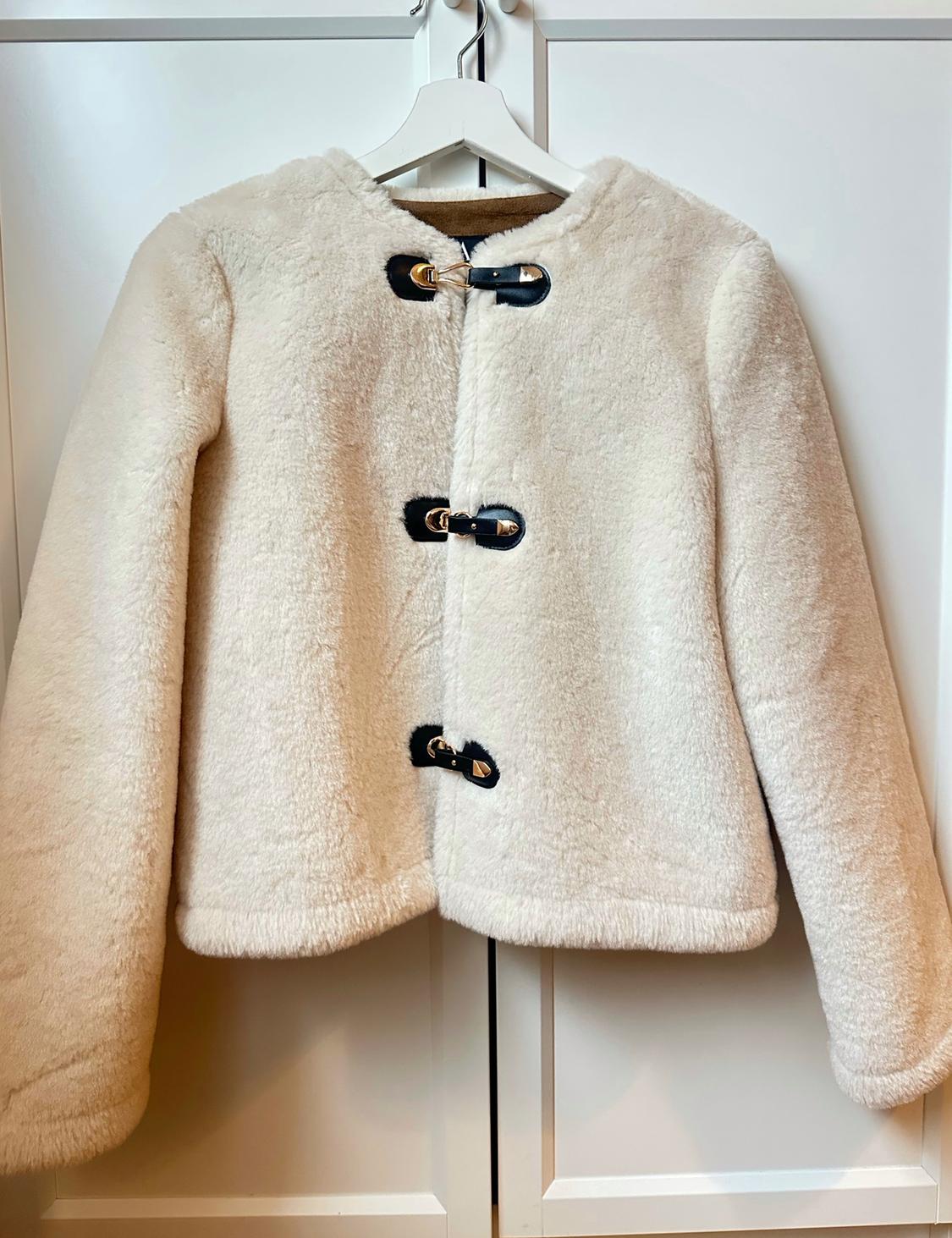 White wool jacket with black buckle fastenings and gold detailing, hanging on a white hanger in front of a wardrobe.