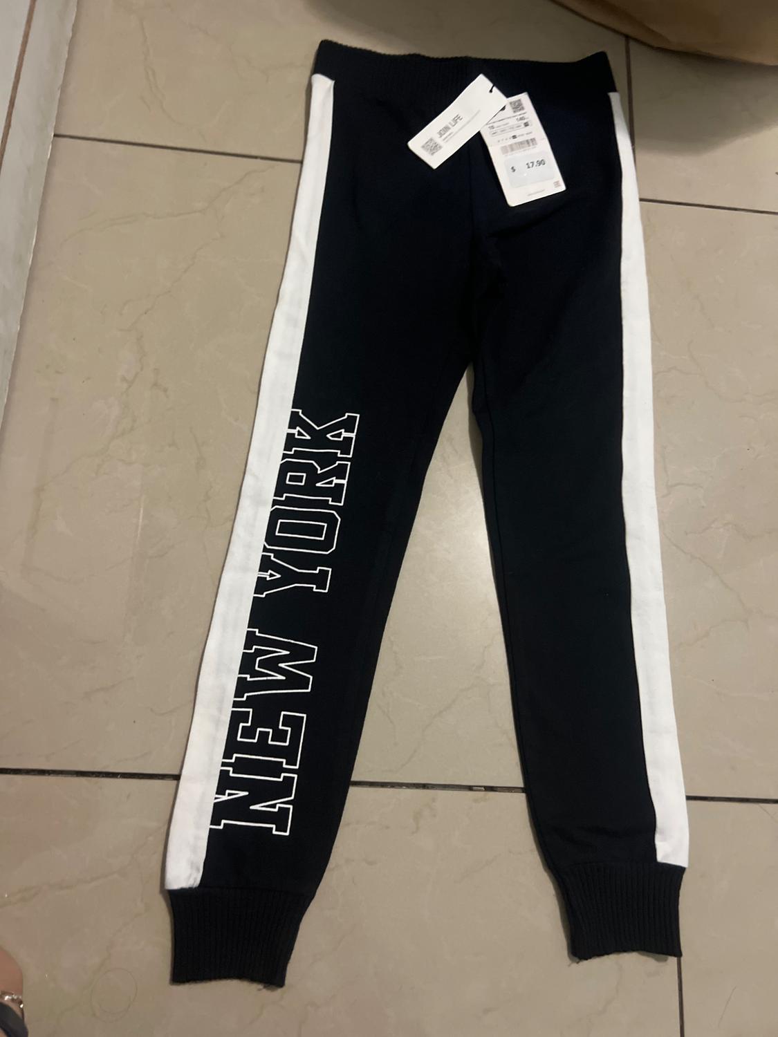 Black trousers with white stripes and "NEW YORK" text on one leg, on a tiled floor. Price tag visible.