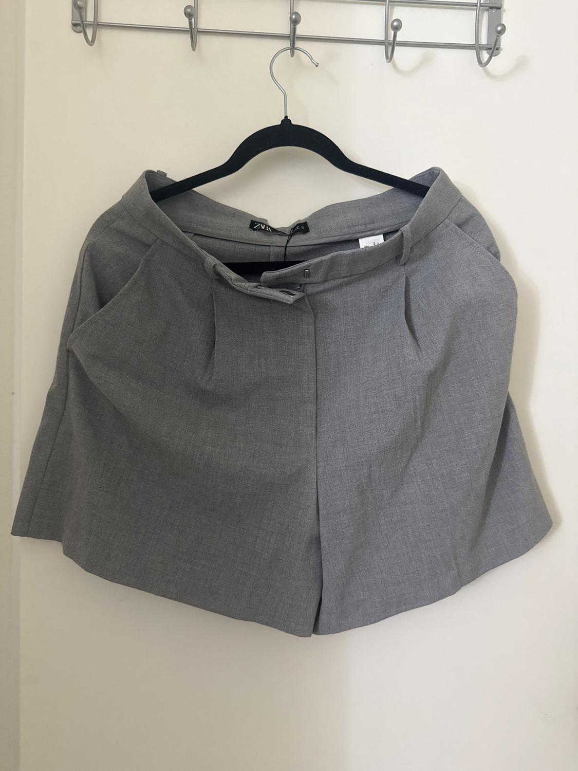 Grey skirt hanging on a black hanger on a metal wall hook. The fabric has a smooth finish and a pleated design.