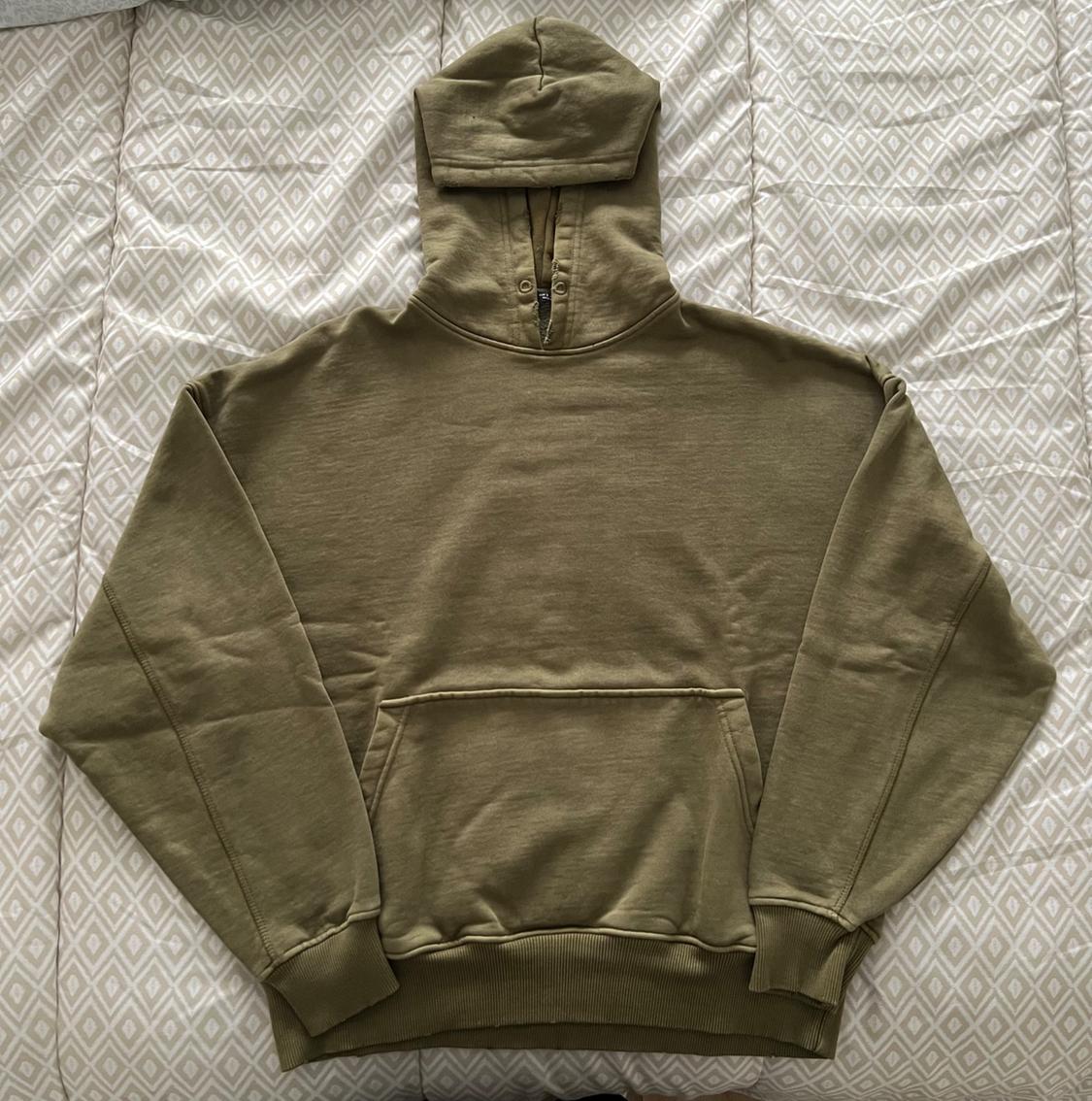 Olive green hoodie on a geometric pattern quilt. It has a front pocket and a neck closure.