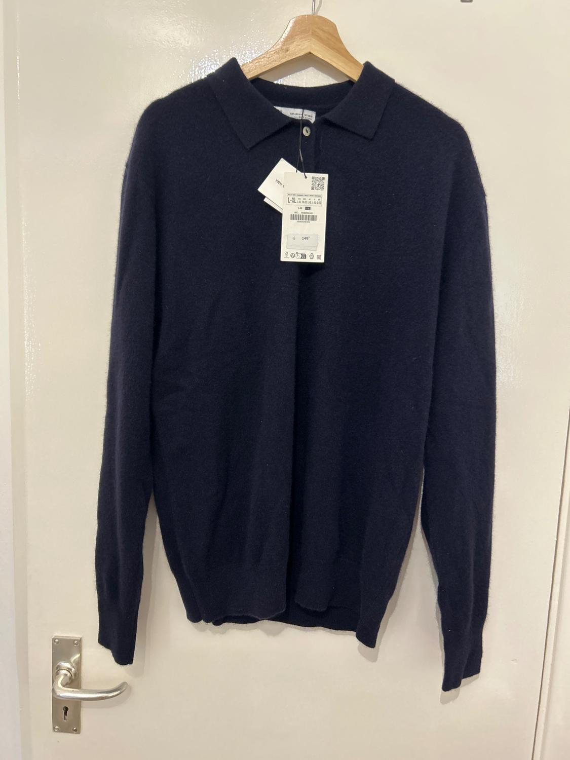 Black long-sleeved polo neck jumper, hanging on a wooden hanger. Visible tag with barcode and price.