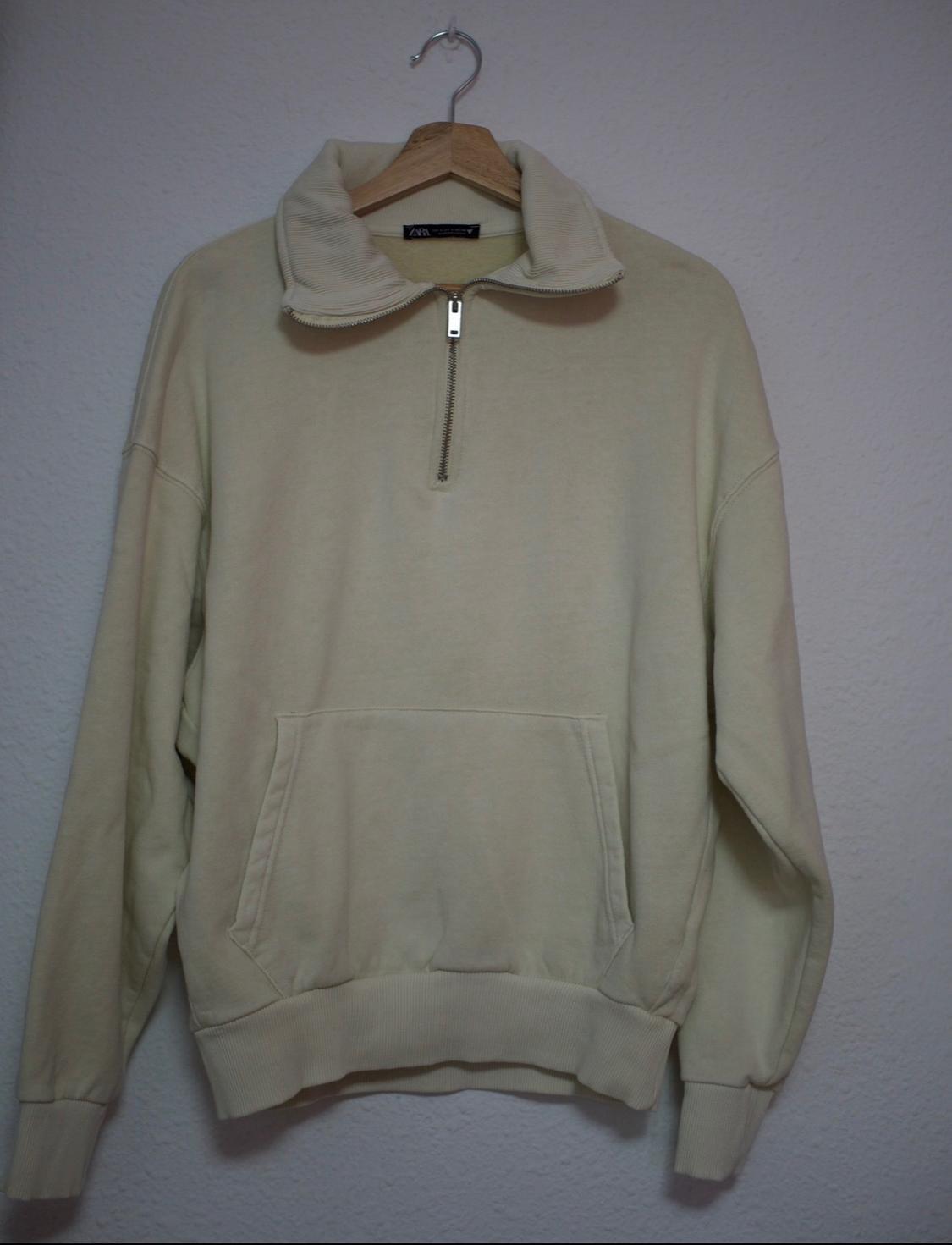 Beige half-zip sweatshirt with a kangaroo pocket and ZARA label. It is hanging on a wooden hanger against a white wall.