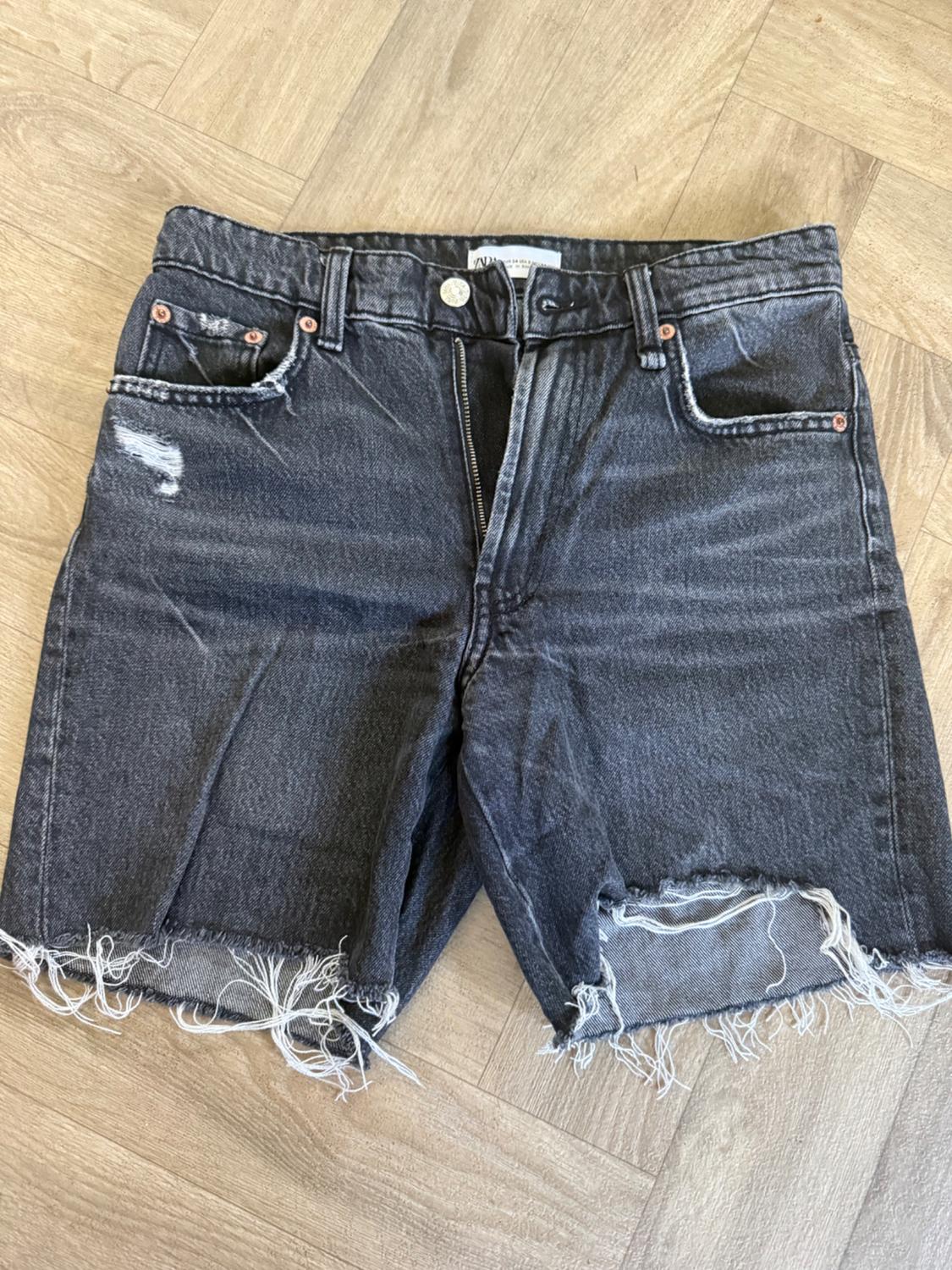 Black distressed denim shorts with rips, frayed hem and open zip on a wooden floor. ZARA label.