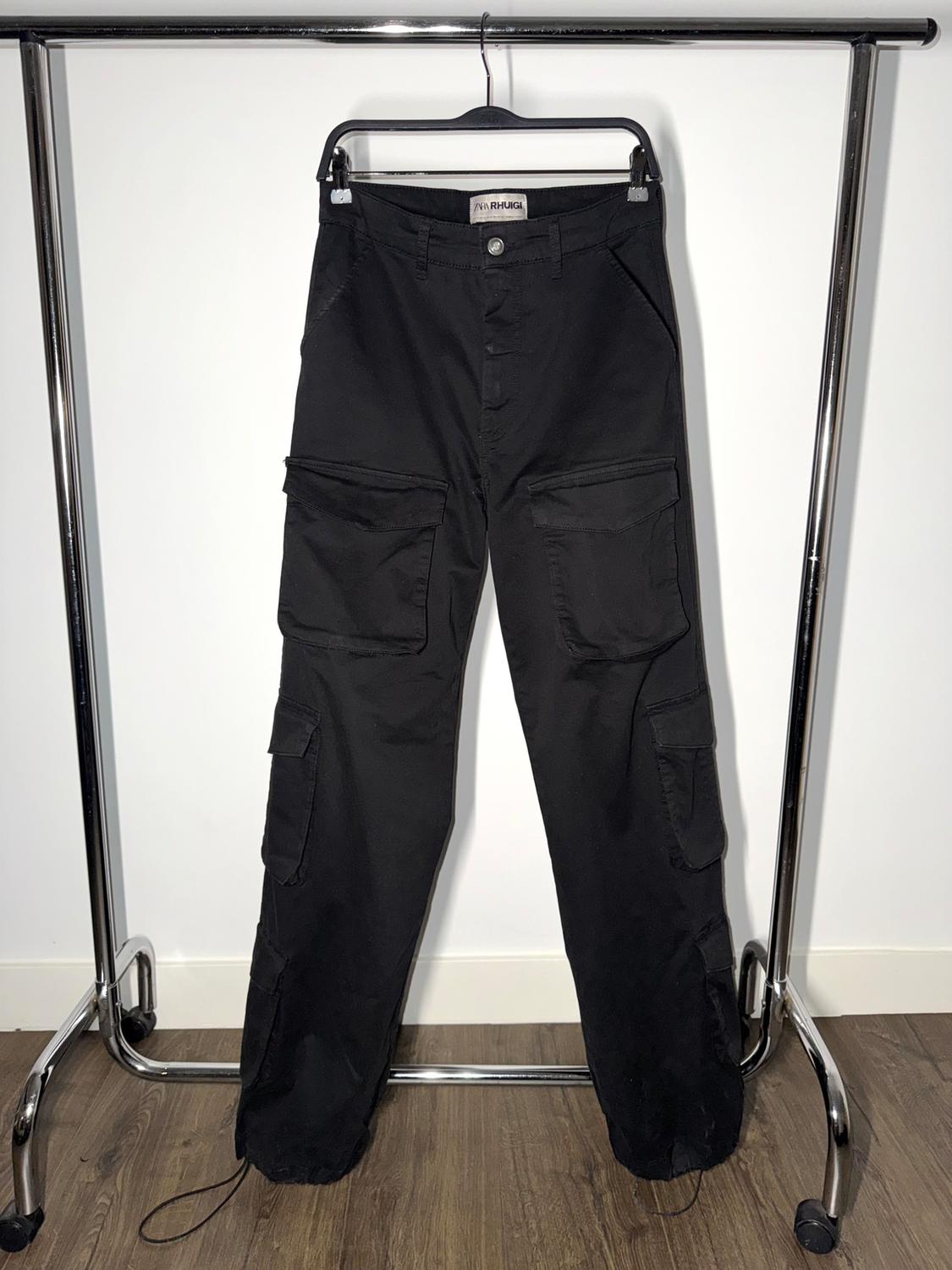 Black cargo trousers hanging on a metal clothes rail. White background and wooden floor. Visible label: "MR RHUGI".