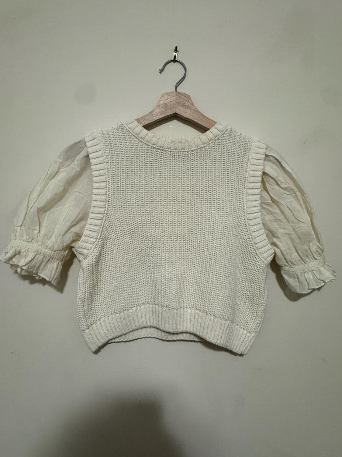 White top on a wooden hanger against a light wall. Knit body with a round neckline and short, ruffled puff sleeves.
