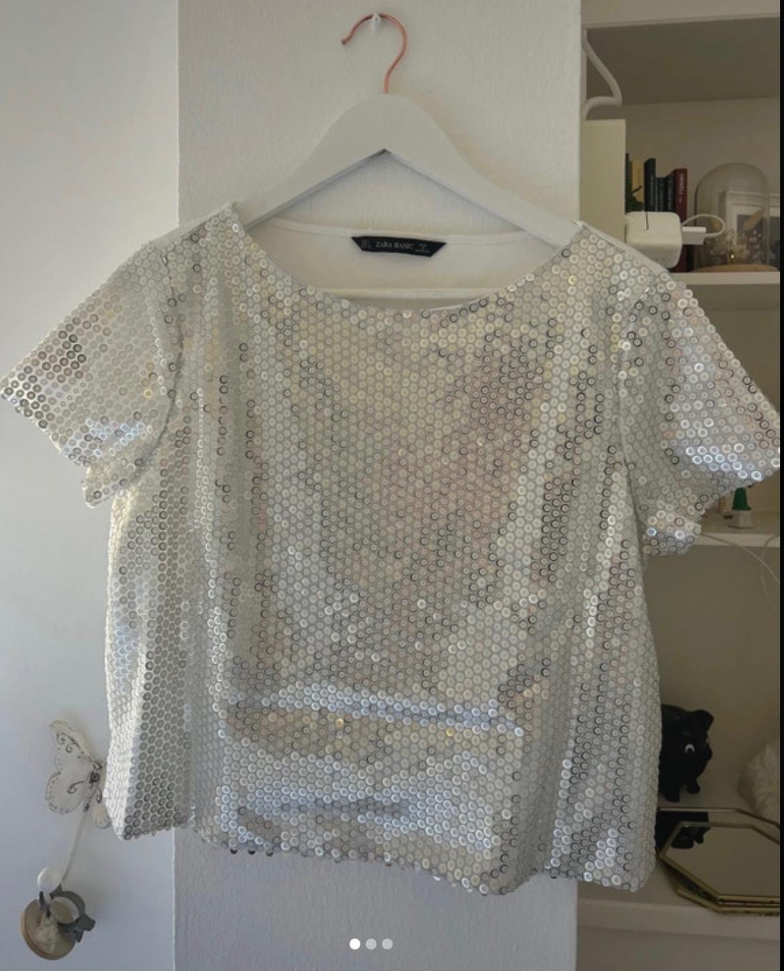 Silver sequined blouse hanging on a white hanger, white wall background and shelf with books and decorative objects.