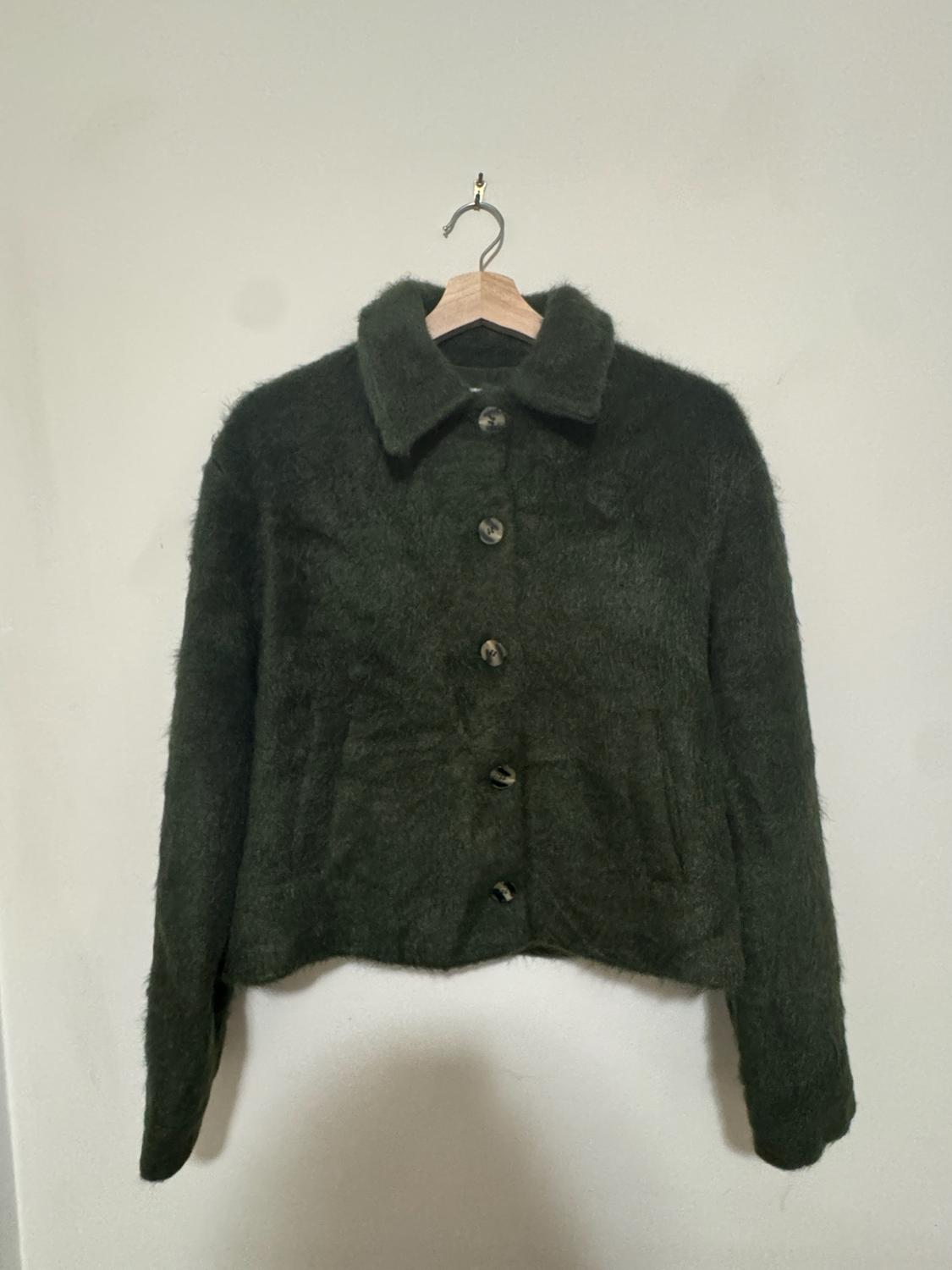 Dark green, soft-textured jacket with a collar and buttons, hanging on a wooden hanger against a light-colored wall. Relaxed style.