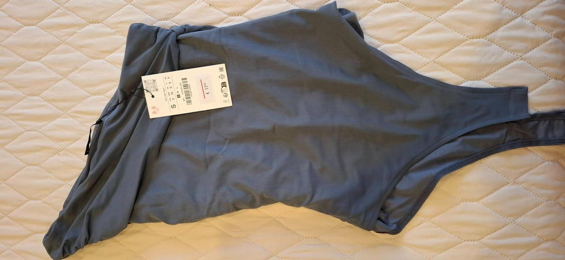 Blue bodysuit with a tag showing the text S and €17.95, on a white quilted surface with a diamond pattern.