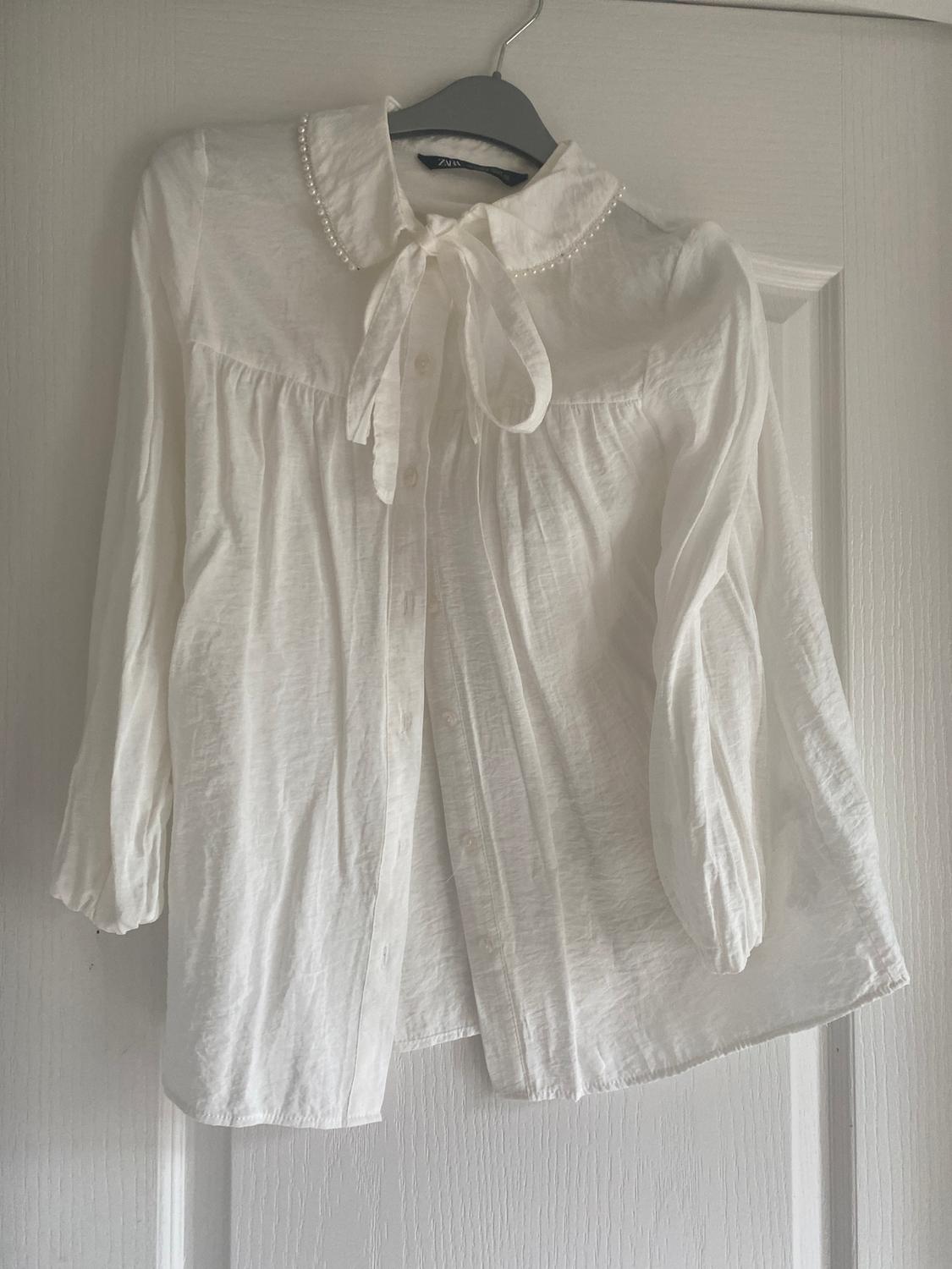 White long-sleeved blouse with a pearl-embellished collar and bow, hanging on a grey hanger over a white door.