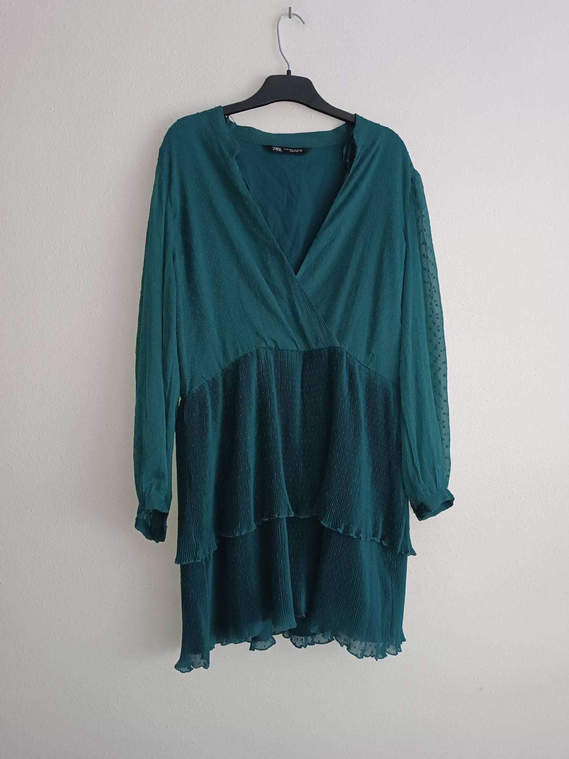 Long-sleeved green dress with a layered design and V-neck, hanging on a black hanger against a white wall.