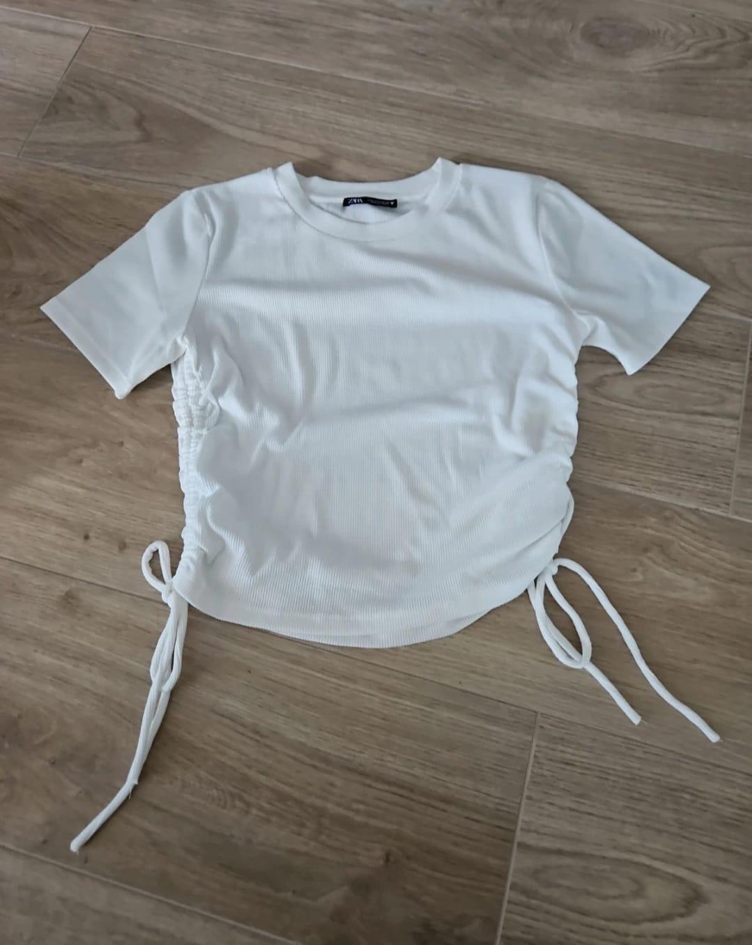 White short-sleeved T-shirt with side ruching and drawstrings, on a light wooden floor.