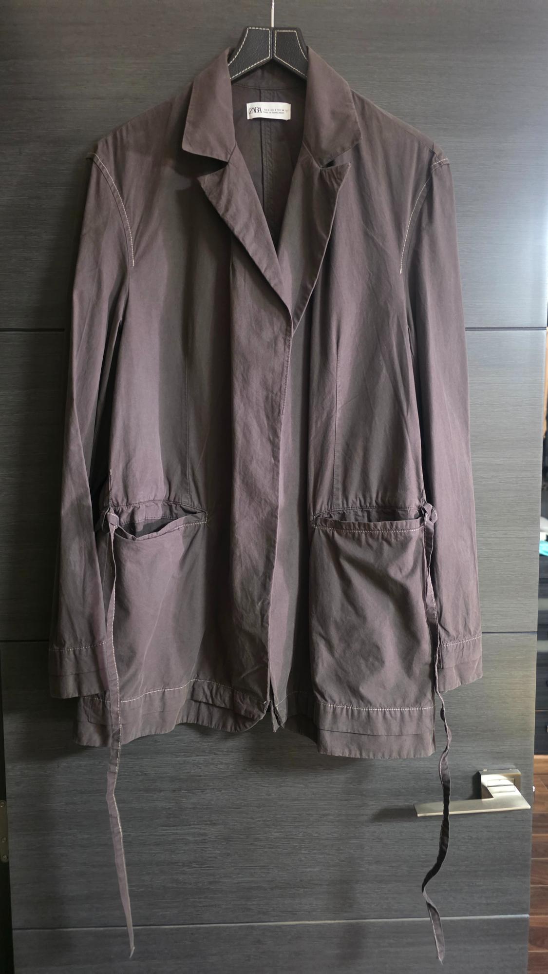 Dark grey jacket with pockets and hanging ribbons, hanging on a dark wood door. Visible label with the text ZARA.