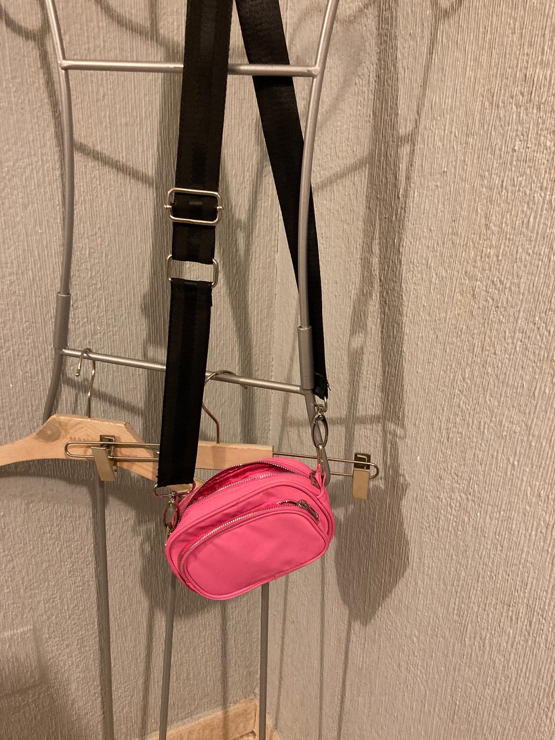 Small pink handbag with zips hanging on a metal coat rack, next to an adjustable black strap. Textured wall background.