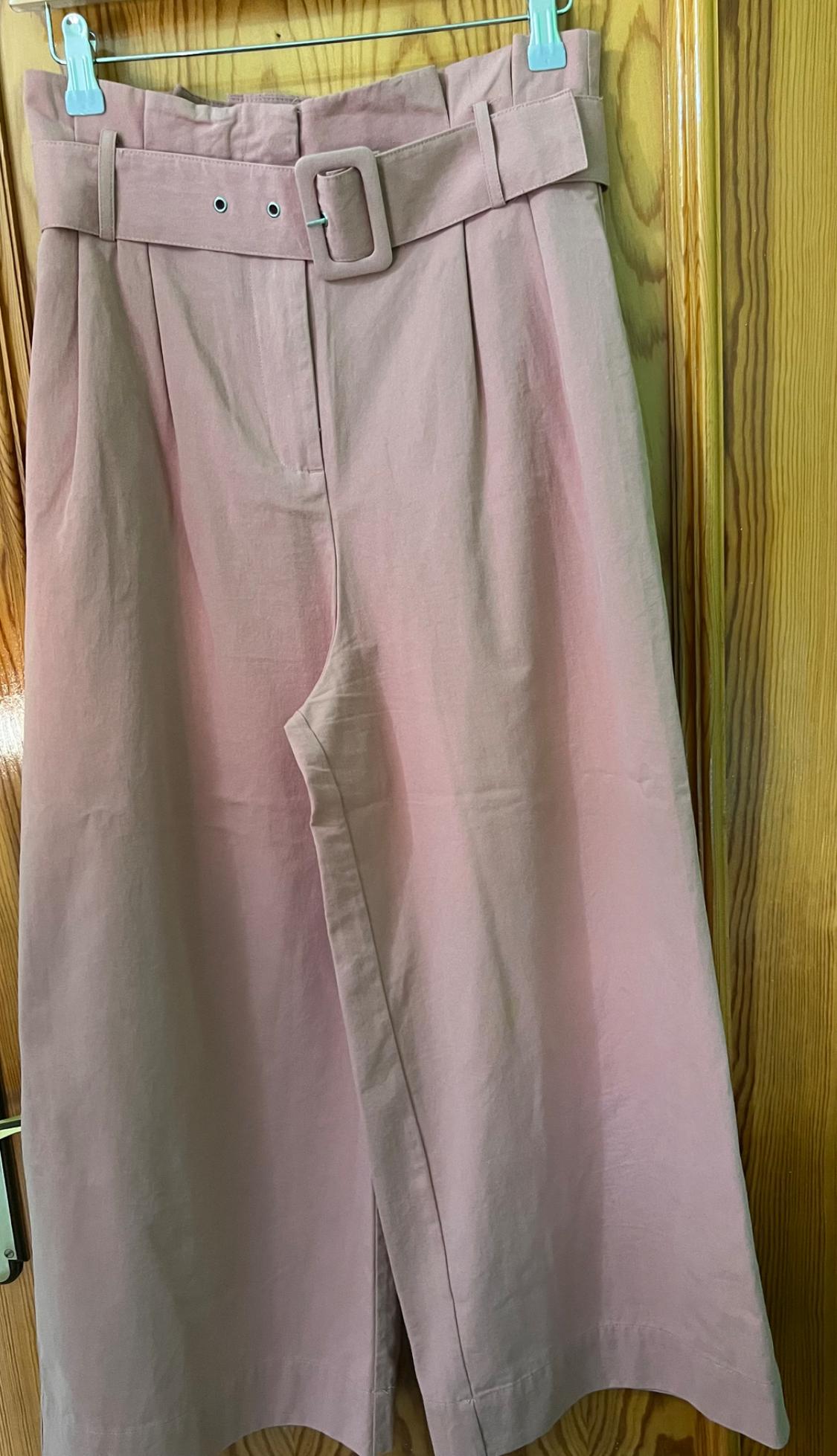 Light pink culotte trousers with a wide belt and square buckle, hanging on a hanger over a wooden door.