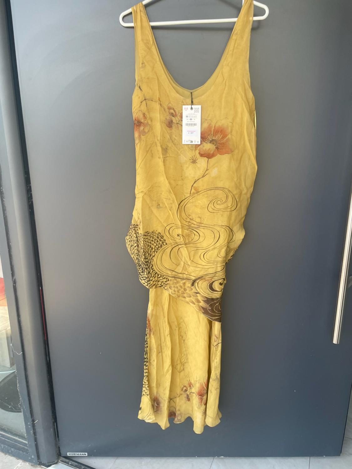 Κίτρινο μακρύ φόρεμα χωρίς μανίκια με φλοράλ στάμπα και κυματιστά τελειώματα, κρεμασμένο σε μια κρεμάστρα σε μια γκρι πόρτα με το κείμενο HORMANN.