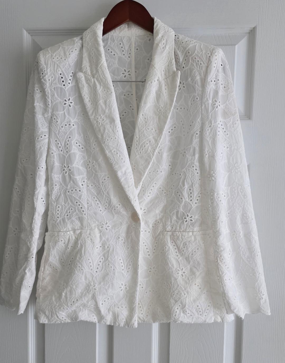 White floral lace blazer hanging on a wooden hanger over a white door.