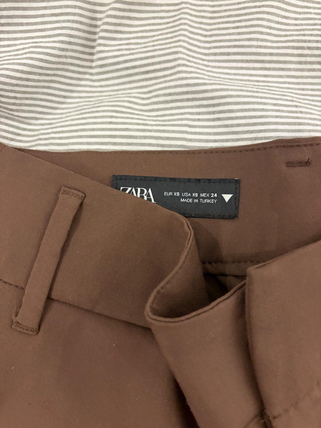 Brown trousers with internal Zara label, size XS, made in Turkey. White and grey striped fabric background.