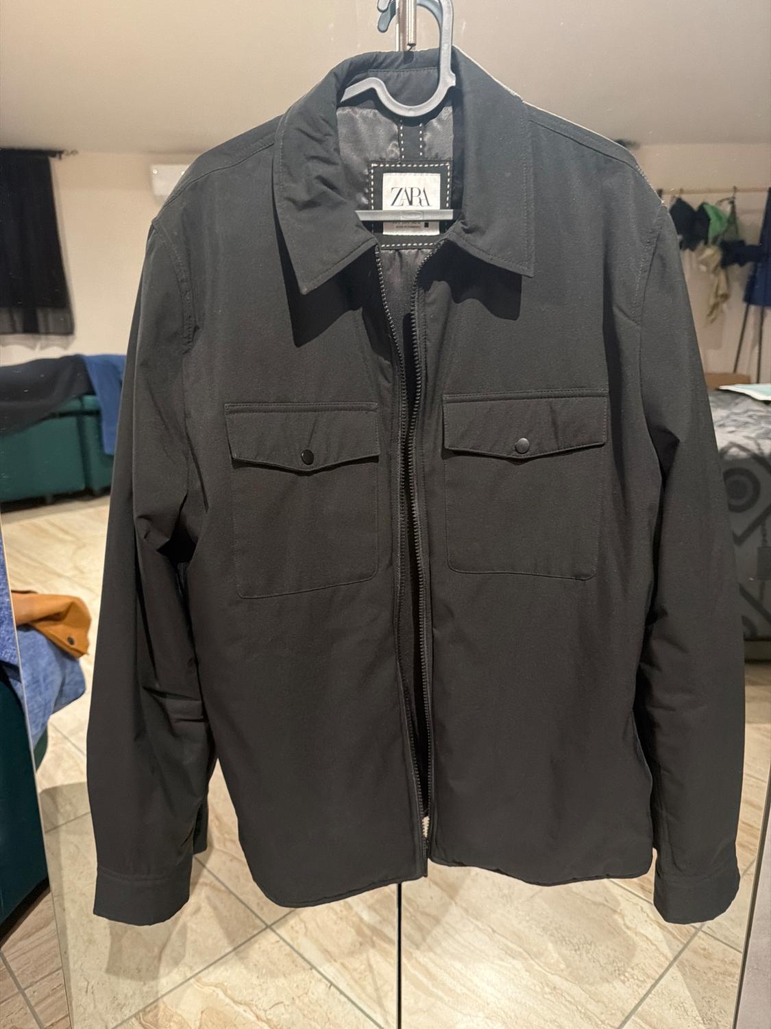 Black jacket with zip and front pockets hanging on a hanger. Visible label with the text "ZARA". Room background.