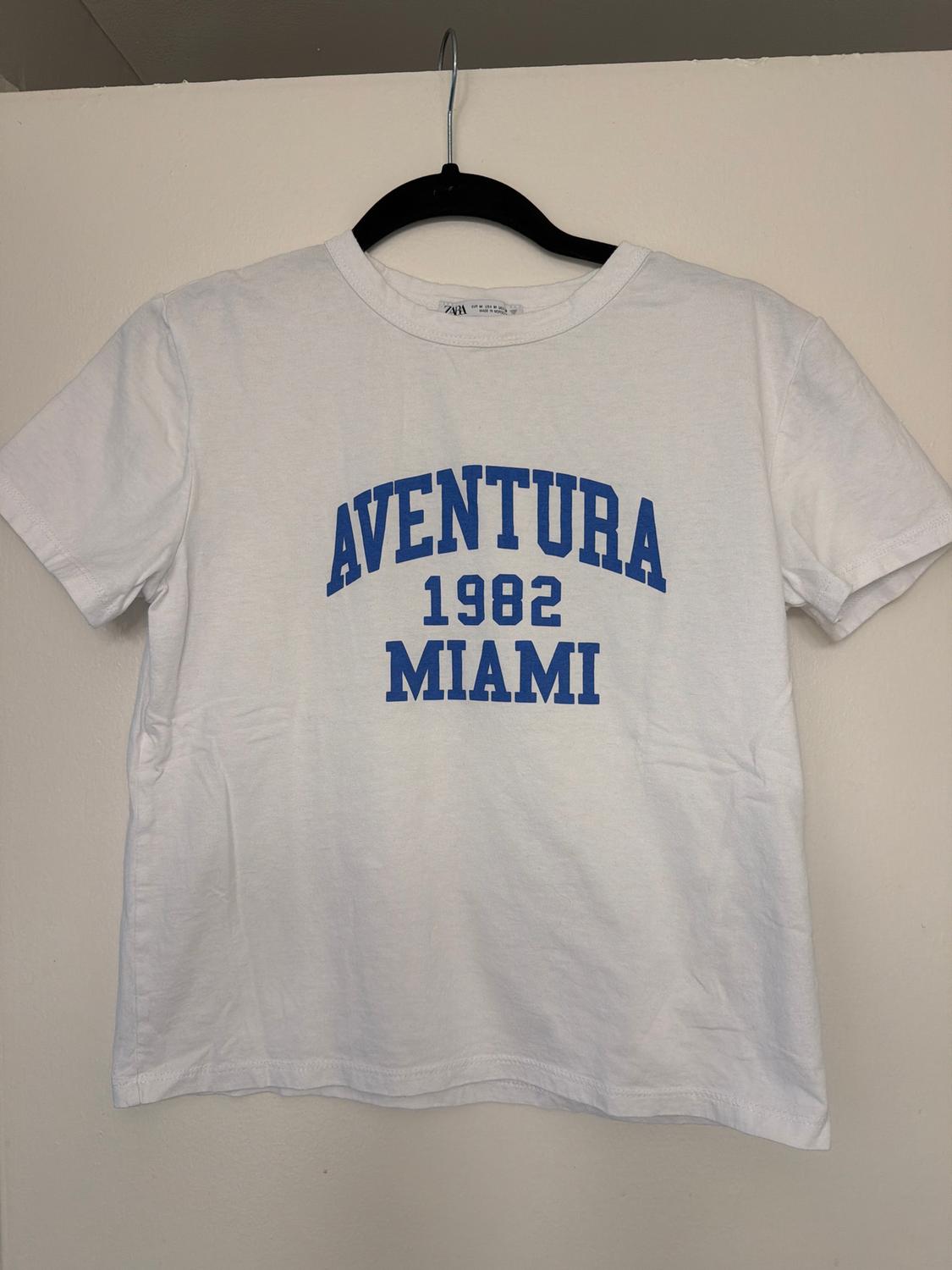 White T-shirt with blue text "AVENTURA 1982 MIAMI" hanging on a black hanger against a white background.