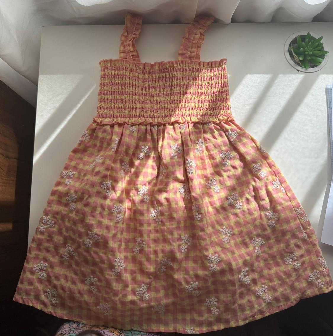 Pink and yellow checked dress with a ruched bodice and embroidered flowers, on a white table with a plant and natural light shadows.