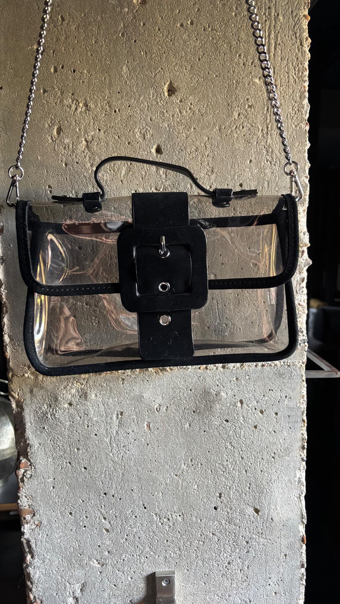 Transparent bag with black trim, short handle, buckle and silver chain, hanging on a textured concrete pillar.