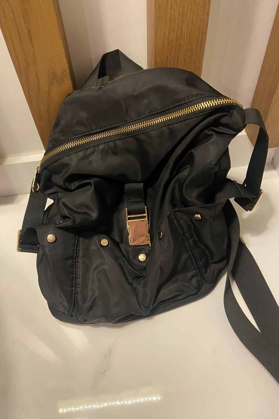 Front view of black backpack with gold zip and metal buckle fastening.
