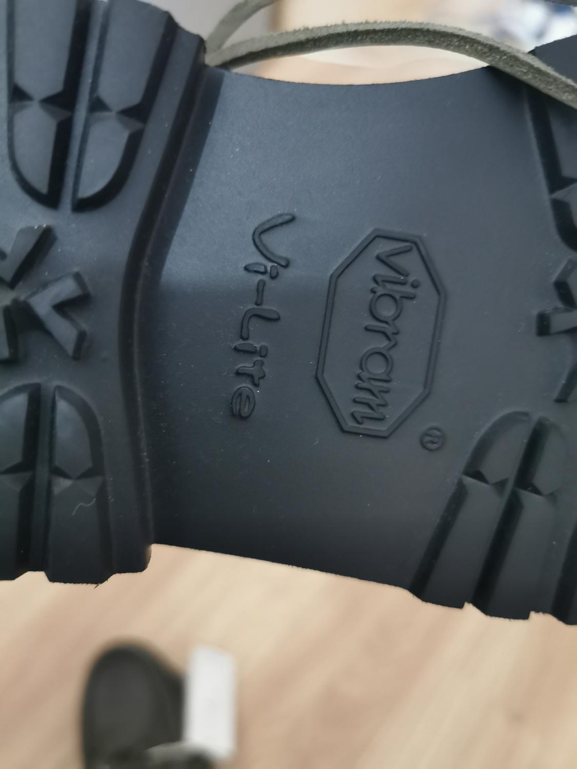Black shoe sole with the text "Vibram Vi-Lite" embossed. Blurred light wood background.