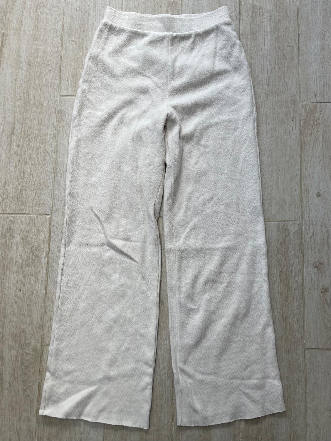 White, soft fabric trousers with a straight leg, elasticated waist and simple design, placed on a light wood floor.