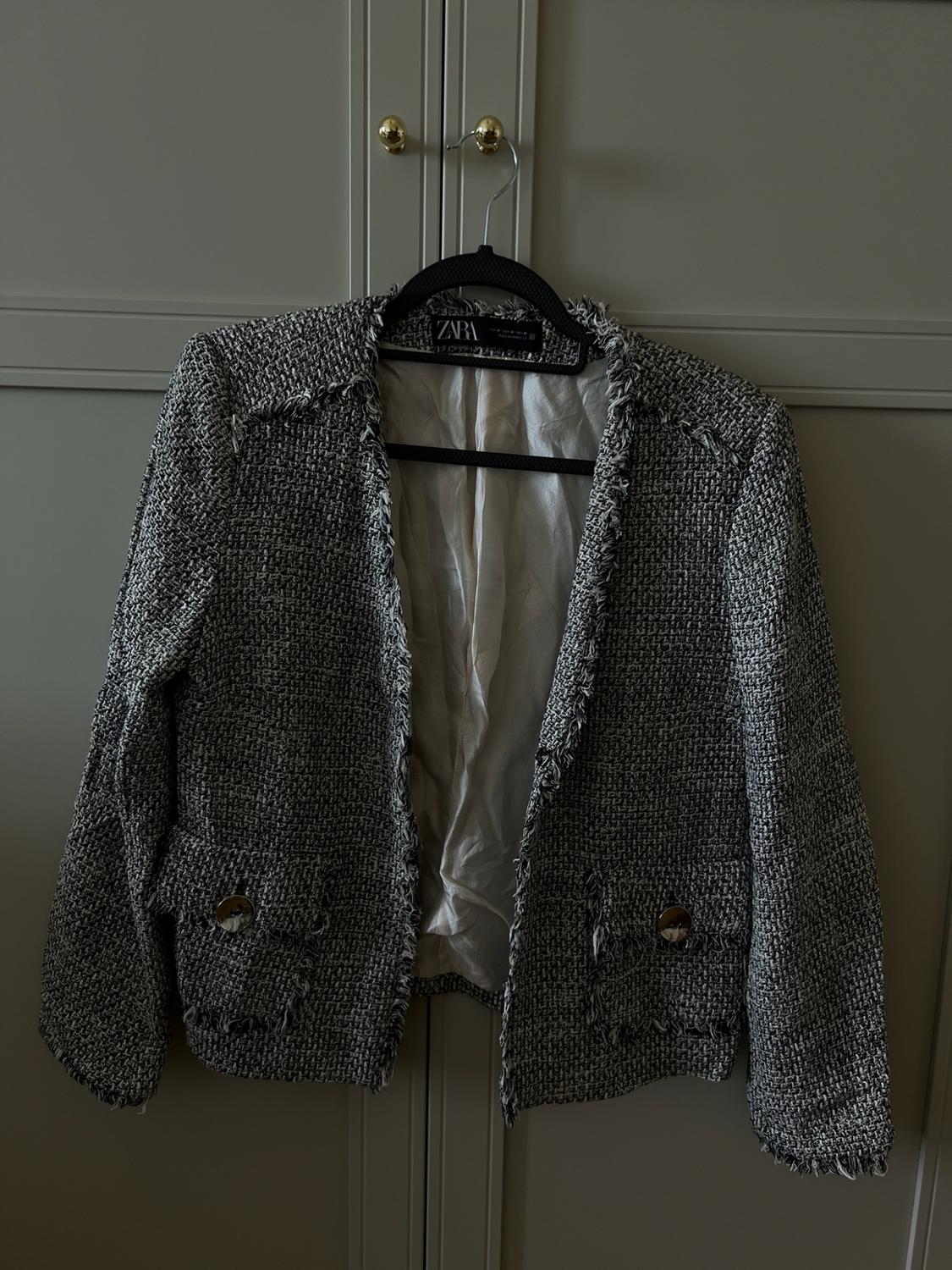 Black and white woven jacket with frayed edges, hanging on a hanger in front of a wardrobe. ZARA label visible on the inside.