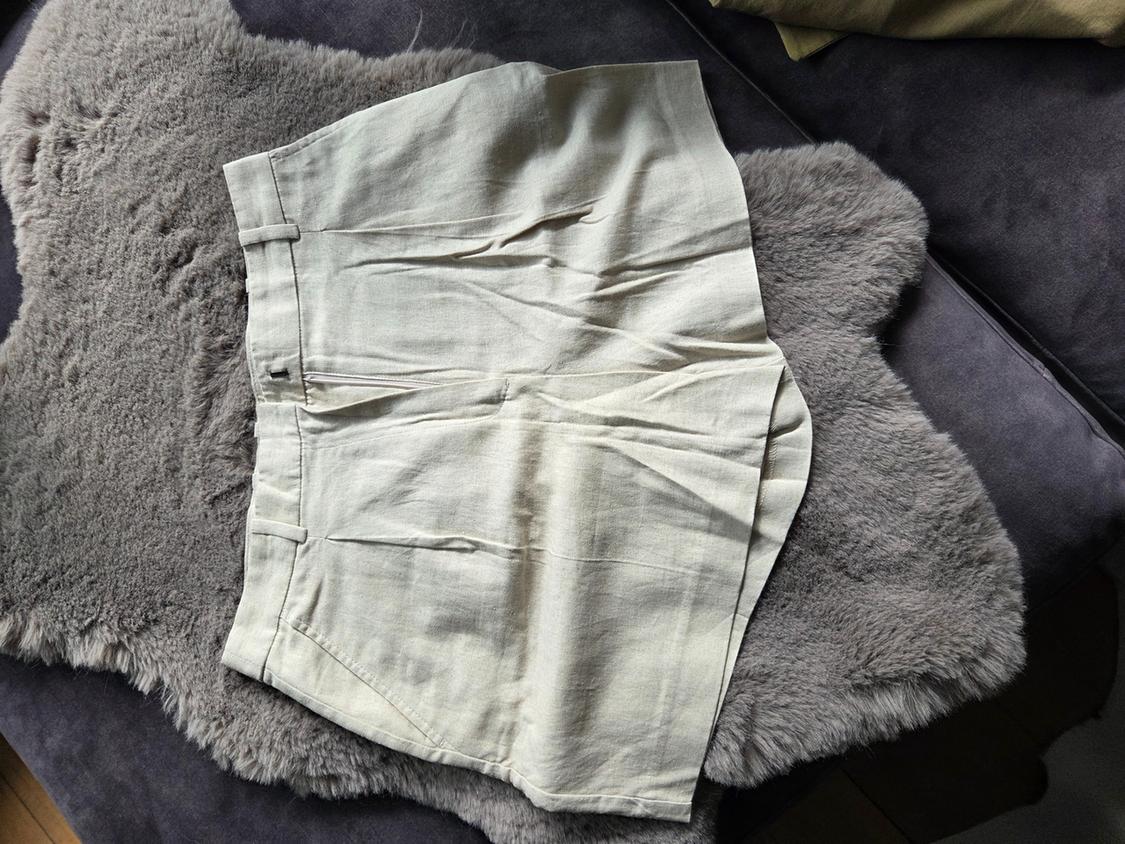 Beige tailored shorts with pleats and belt loops, displayed on a grey shaggy rug and a dark sofa.